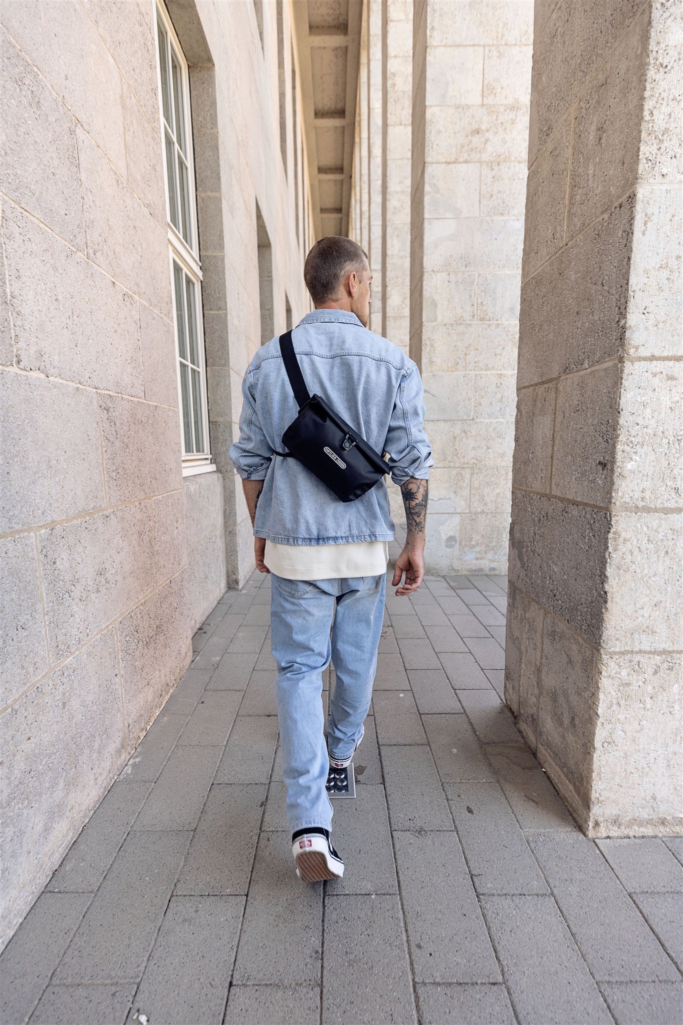 Vue arrière d’un homme avec sac bandoulière noir ORTLIEB Velo-Sling Flex à fermeture roulée marchant sous une colonnade.