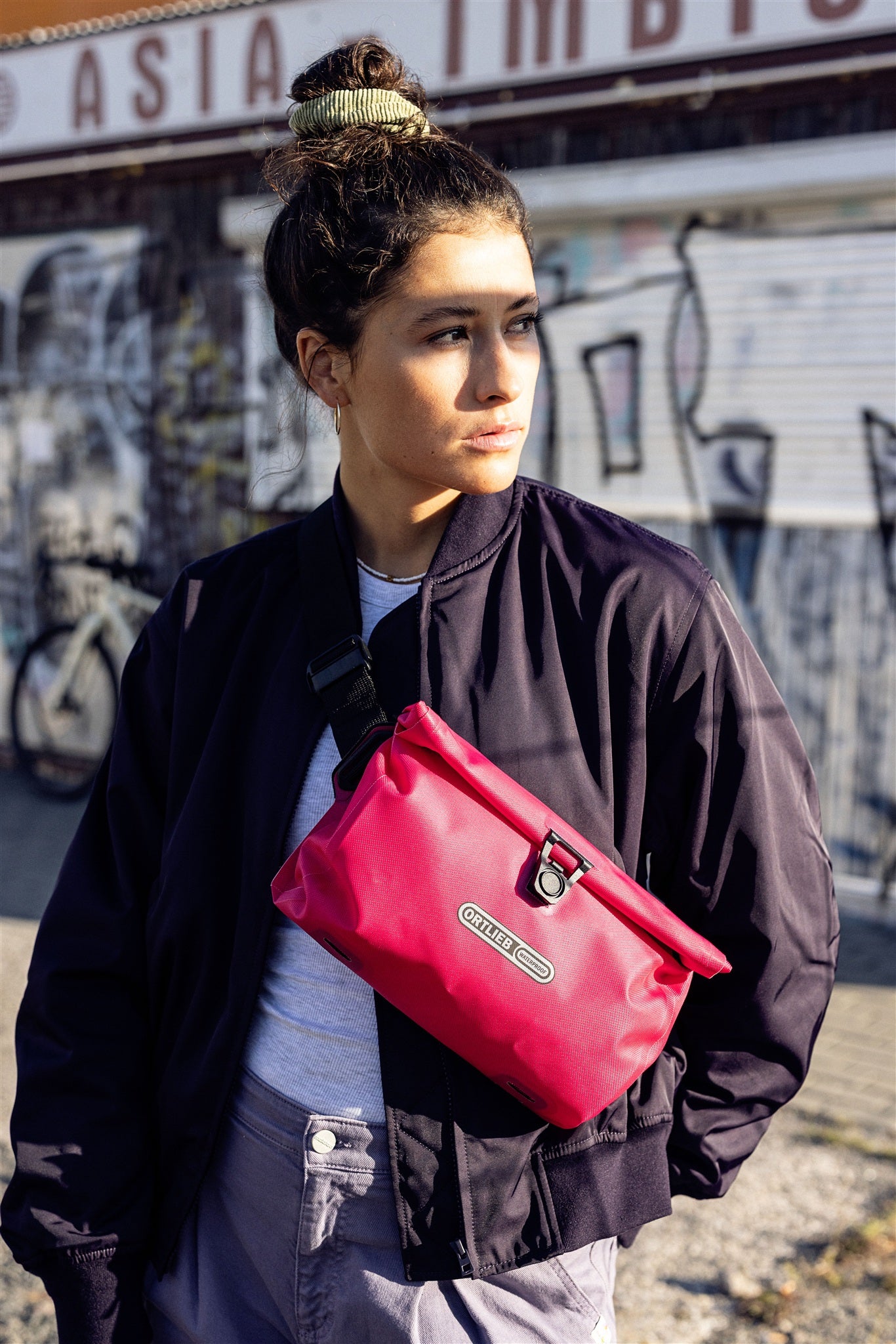 Portrait d’une femme avec sac bandoulière ORTLIEB Velo-Sling Flex couleur cyber-pink dans un décor urbain.